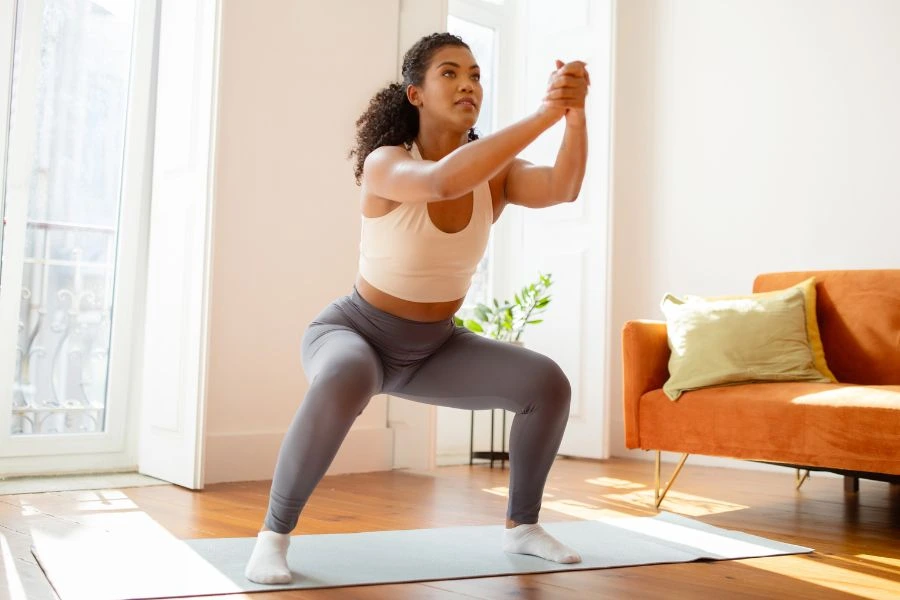 Mulher jovem realizando agachamento em um tapete de yoga, usando top esportivo bege e legging cinza, em um ambiente iluminado por luz natural. A sala possui janelas amplas, um sofá laranja com almofada verde e uma pequena planta decorativa, criando um espaço caseiro acolhedor. A postura mostra foco em exercícios físicos e bem-estar.