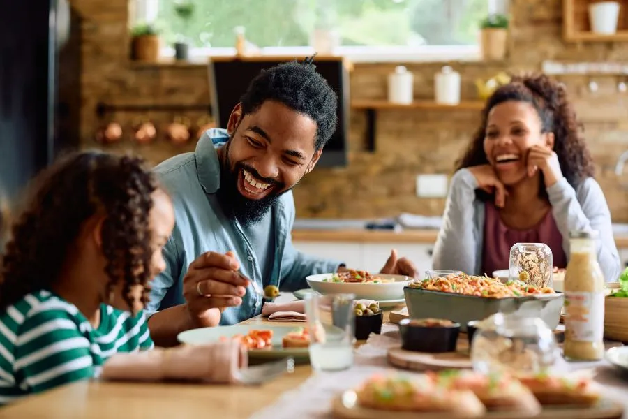 Família composta por pai, mãe e filha compartilhando uma refeição saudável em um ambiente de cozinha aconchegante e iluminado. A cena mostra sorrisos e interação alegre enquanto pratos com alimentos frescos, como saladas, pães e legumes, estão dispostos sobre a mesa de madeira. Ao fundo, detalhes de uma cozinha rústica com prateleiras decorativas, criando um clima acolhedor e familiar.