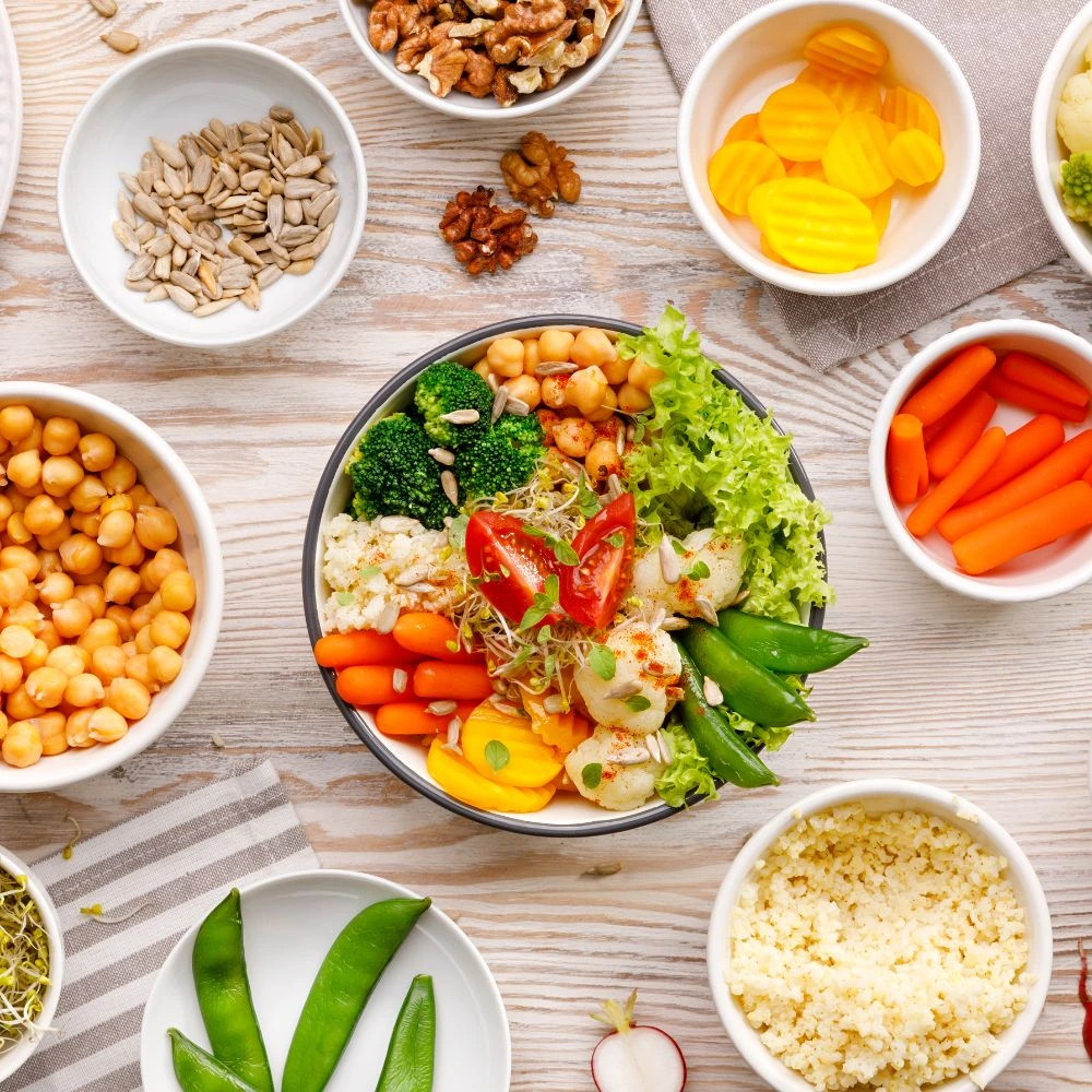 Tigela de salada vibrante composta por legumes frescos, como brócolis, alface, tomate-cereja, cenoura, ervilhas e beterraba em rodelas, acompanhada de grãos como grão-de-bico e quinoa. A mesa está decorada com pequenos bowls de nozes, sementes de girassol e vegetais crus como cenouras e ervilhas-tortas, criando um cenário colorido e saudável, ideal para promover alimentação equilibrada baseada em ingredientes naturais.