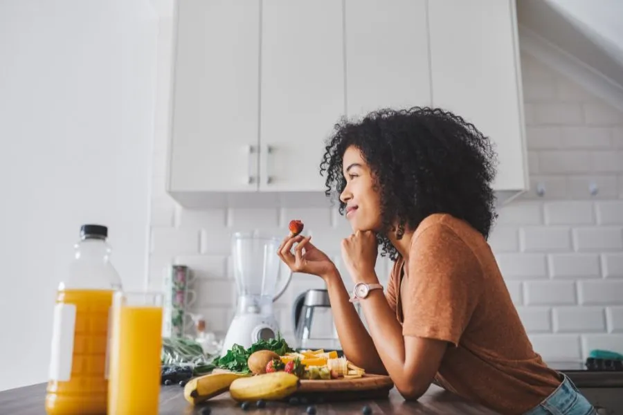 Mulher jovem com cabelo cacheado, vestindo uma camiseta marrom, segurando um morango enquanto sorri levemente em uma cozinha moderna com parede de azulejos brancos. Em uma mesa de madeira à sua frente, há garrafas de suco de laranja, frutas frescas como bananas, maçãs e mirtilos, além de uma tábua com alimentos, transmitindo um momento saudável e descontraído.