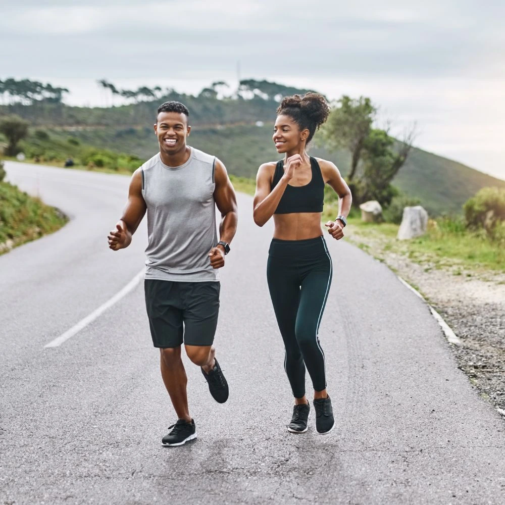 Homem e mulher jovens correndo juntos ao ar livre em uma estrada asfaltada cercada por uma paisagem natural, com colinas verdes e árvores ao fundo. O homem veste camiseta cinza sem mangas, shorts pretos e tênis escuros, enquanto a mulher usa um top esportivo preto, legging preta com detalhes laterais e tênis pretos. Ambos sorriem, transmitindo energia, saúde e a prática de exercícios físicos ao ar livre em um dia nublado.