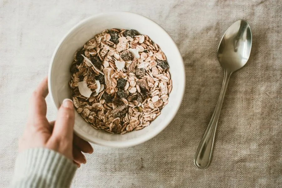 Tigela branca com uma mistura de granola caseira, contendo aveia, passas, lascas de coco e outros ingredientes naturais, segurada por uma mão sobre uma toalha de linho bege. Ao lado da tigela, uma colher de aço inoxidável compõe um ambiente convidativo e saudável para o café da manhã.