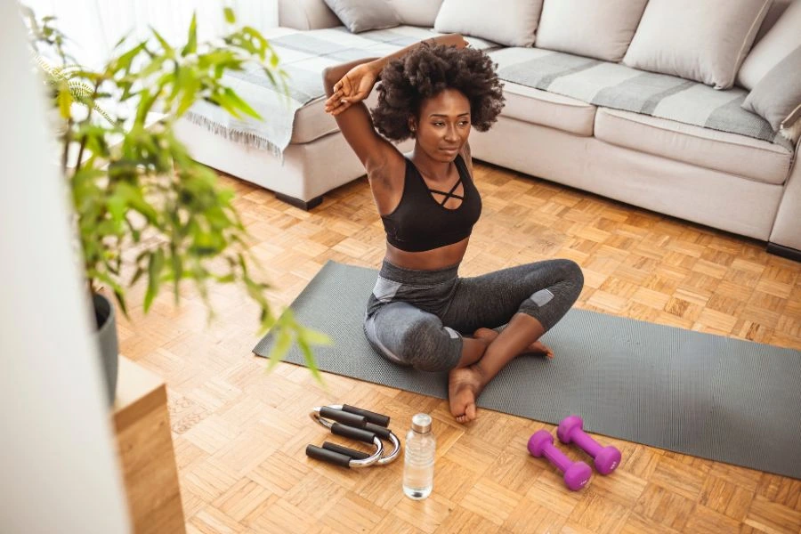 Mulher jovem praticando alongamento em casa, sentada em um tapete de yoga. Ela usa roupas esportivas pretas e está em um ambiente bem iluminado por luz natural, com sofás claros ao fundo e plantas decorativas. Pesos roxos, uma garrafa de água e um corda de pular estão organizados ao lado, simbolizando uma rotina de exercícios e bem-estar em um espaço doméstico confortável.
