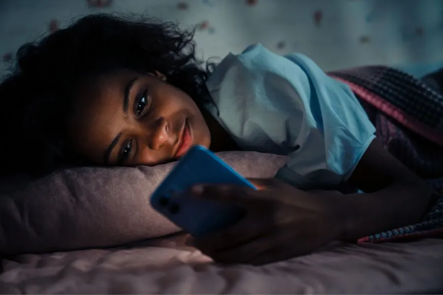 Jovem sorridente usando celular na cama durante a noite em ambiente escuro, ilustrando hábitos digitais noturnos e possíveis impactos na qualidade do sono e saúde mental