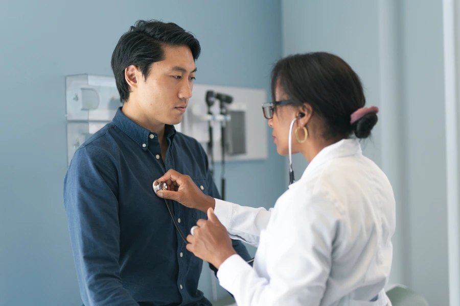 Médica utilizando estetoscópio para examinar o tórax de um paciente sentado, que veste camisa azul. O consultório ao fundo apresenta paredes claras, instrumentos médicos e iluminação suave, transmitindo um ambiente profissional e de cuidado com a saúde.