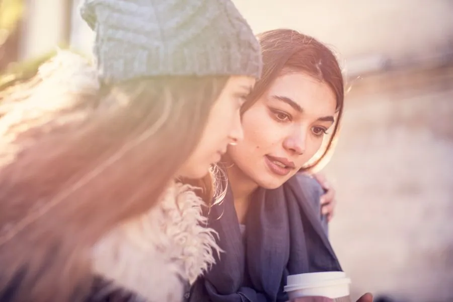 Duas mulheres em um momento de conversa e apoio emocional ao ar livre. Uma delas usa gorro cinza e casaco com pelos, enquanto a outra, de cabelos soltos e enrolada em um lenço azul, segura um copo descartável. Cena iluminada pela luz suave do sol, transmitindo conforto e empatia.