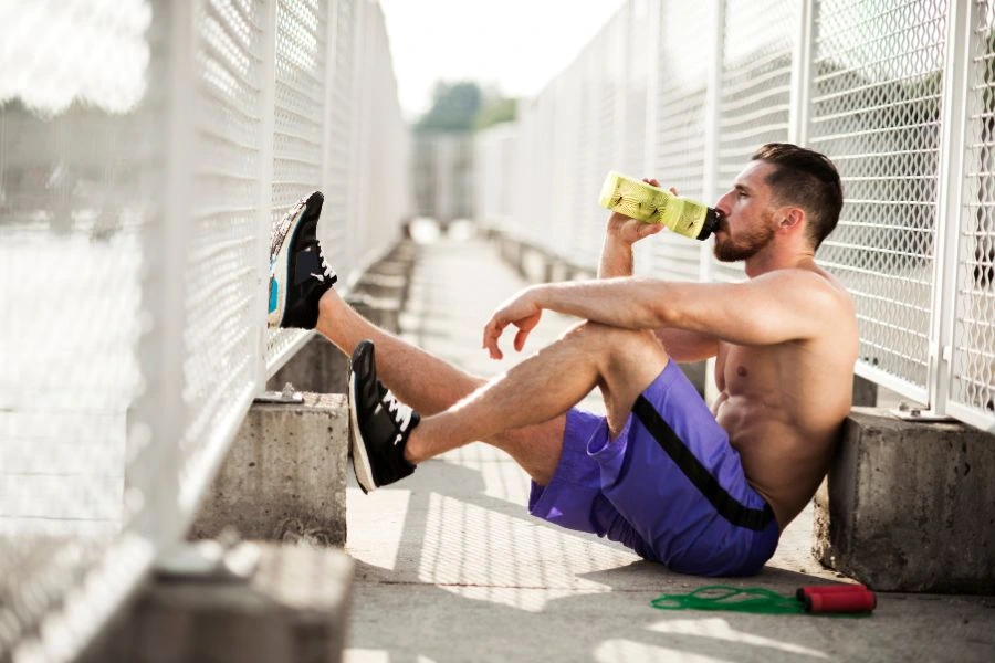 Homem jovem descansando sentado no chão de uma ponte cercada por grades, vestindo shorts roxos e tênis, enquanto bebe água de uma garrafa esportiva verde. Ao lado dele, um elástico de treino está visível, sugerindo uma pausa após um treino ao ar livre em um dia ensolarado. Ambiente transmite estilo de vida ativo e foco em saúde e bem-estar.