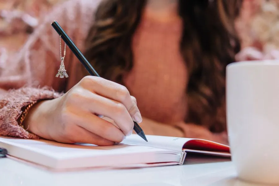Close de uma mão feminina escrevendo em um caderno com capa branca e vermelha, segurando uma caneta preta decorada com um pingente da Torre Eiffel. A mulher veste um suéter rosa e há uma caneca branca na mesa ao lado, compondo um cenário aconchegante e criativo.