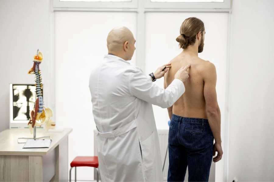 Médico vestindo jaleco branco examinando as costas de um paciente sem camisa em um consultório clínico iluminado. Ao fundo, uma mesa com modelo anatômico da coluna vertebral e radiografias, compondo um ambiente profissional voltado para cuidados com a saúde e ortopedia.
