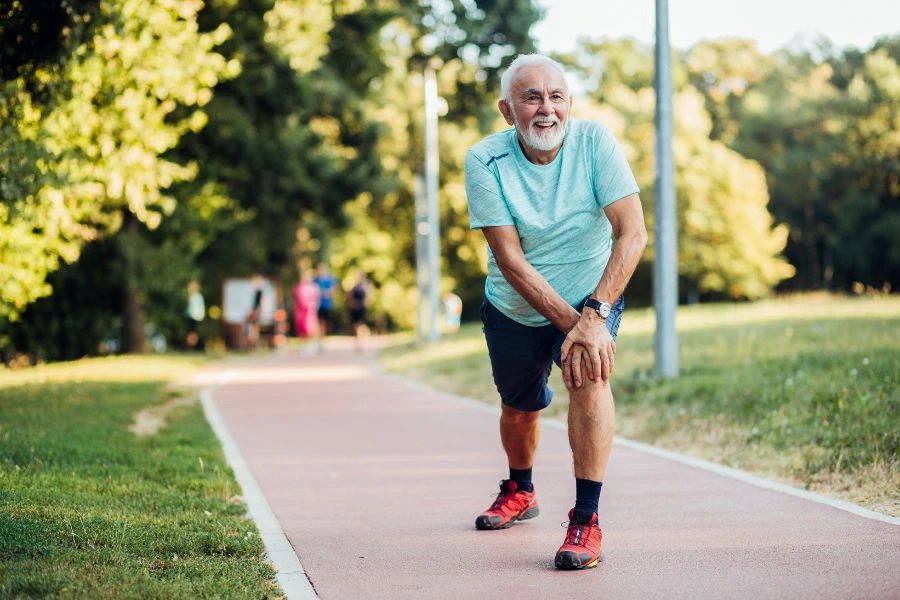 Homem idoso sorridente se alongando ao ar livre em uma pista de caminhada cercada por árvores em um parque. Ele veste uma camiseta azul, shorts escuros e tênis vermelhos, representando hábitos saudáveis, bem-estar e exercícios físicos na terceira idade.