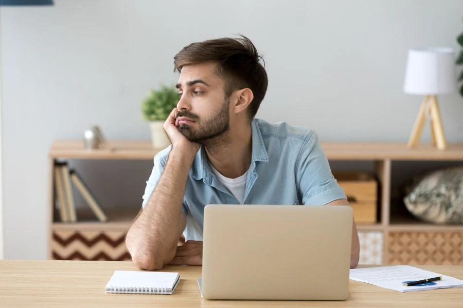 Homem jovem aparentando tédio ou falta de motivação sentado em uma mesa de escritório, apoiando o rosto na mão enquanto olha para o lado. Em frente a ele, estão um laptop, bloco de notas e caneta. Ambiente de trabalho com decoração simples ao fundo, transmitindo um momento de distração ou procrastinação no cotidiano profissional.