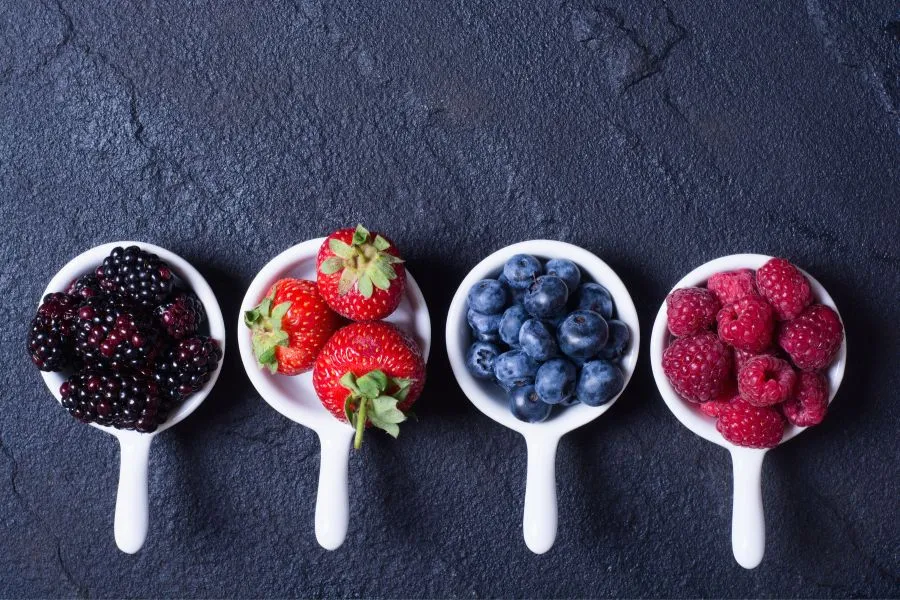 Quatro colheres de cerâmica branca alinhadas sobre uma superfície de pedra escura, cada uma preenchida com frutas frescas: amoras, morangos, mirtilos e framboesas. As cores vibrantes dos frutos contrastam com o fundo escuro, criando um visual atraente que destaca alimentos ricos em antioxidantes e saudáveis.