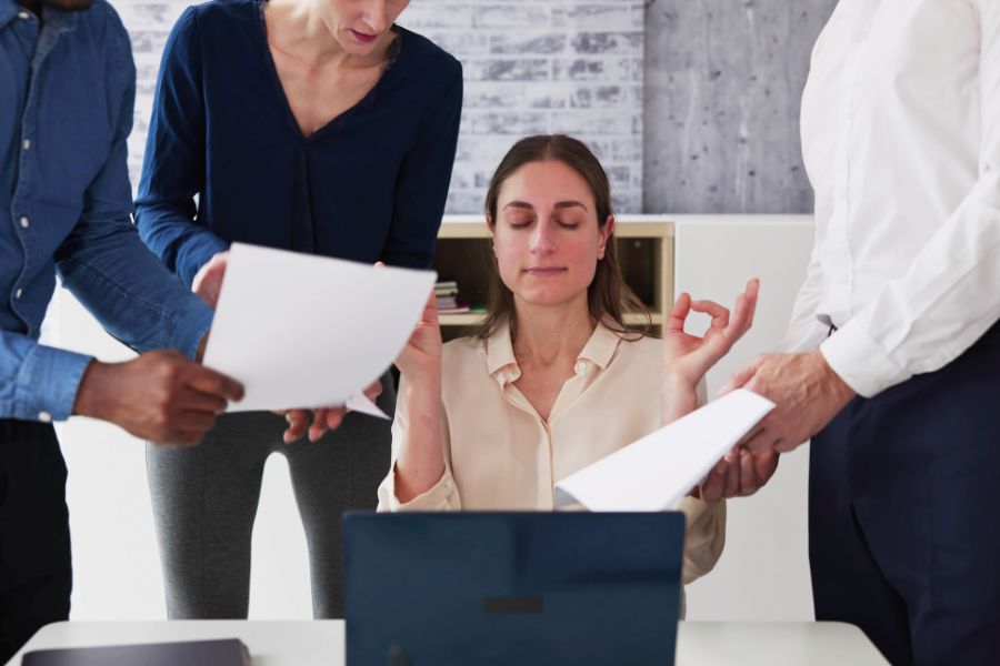 Mulher sentada em frente a um laptop em um escritório, com os olhos fechados enquanto pratica meditação e faz gesto de calma com as mãos em meio a colegas que lhe entregam documentos. Cena representa gerenciamento de estresse, mindfulness e equilíbrio no ambiente de trabalho.