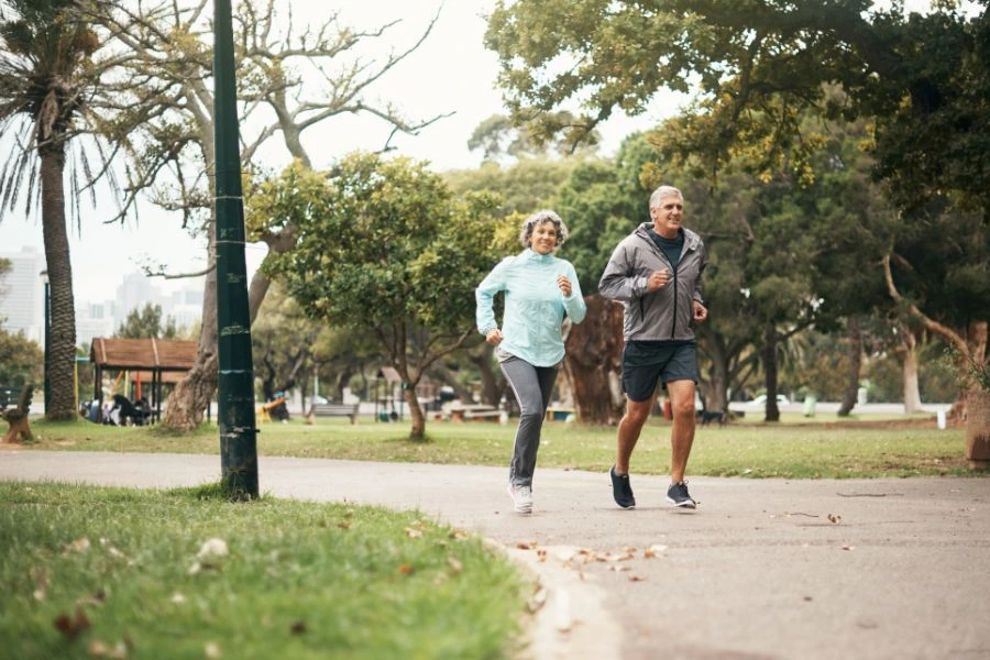 Casal idoso praticando corrida ao ar livre em um parque arborizado em um dia claro. Ambos estão sorrindo enquanto usam roupas esportivas, simbolizando um estilo de vida saudável, bem-estar e atividade física na terceira idade.