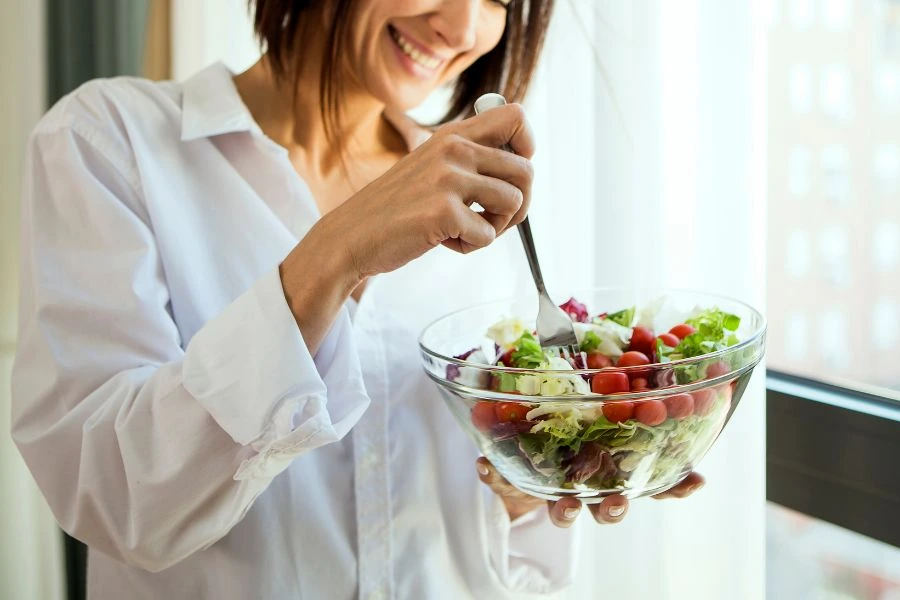 Mulher sorridente segurando uma tigela de vidro com salada fresca, contendo alface, tomates-cereja e vegetais coloridos, misturando os ingredientes com um garfo. Imagem iluminada por luz natural próxima a uma janela, promovendo alimentação saudável e leveza.