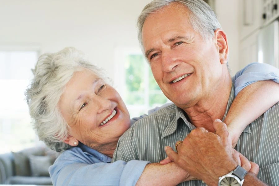 Casal idoso sorridente em um momento de afeto e cumplicidade, com a mulher abraçando o homem por trás enquanto estão em um ambiente claro e acolhedor. Cena representa amor, felicidade e qualidade de vida na terceira idade.