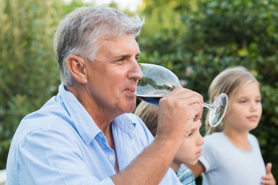 Homem idoso de cabelos grisalhos apreciando um copo de vinho tinto em um ambiente ao ar livre com duas crianças ao fundo. Ele veste uma camisa azul e está cercado por um cenário natural de árvores e plantas, simbolizando momentos de convivência familiar e lazer.