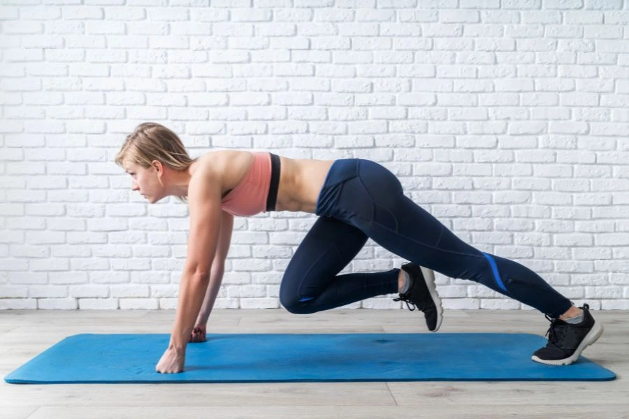 Mulher jovem realizando o exercício 'alpinista' em um tapete de yoga azul, vestindo top esportivo rosa e calça legging azul-marinho, em frente a uma parede de tijolos brancos. O ambiente é bem iluminado, e a postura dela revela foco e força, destacando uma rotina de treino funcional e saúde física.
