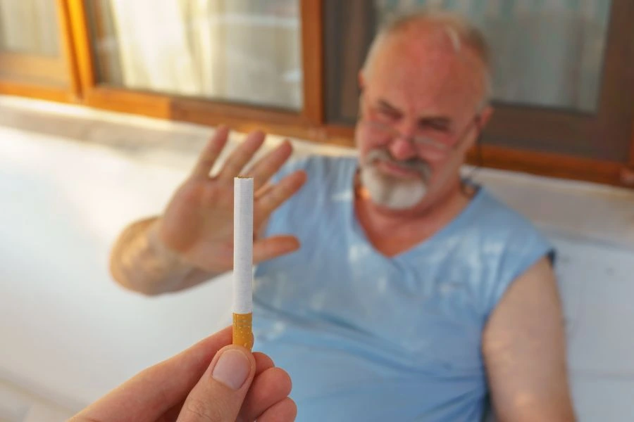 Homem idoso de barba branca sentado e fazendo um gesto de recusa com a mão enquanto alguém oferece um cigarro. Cena representa rejeição ao tabagismo, escolha por uma vida saudável e conscientização sobre os malefícios do cigarro.