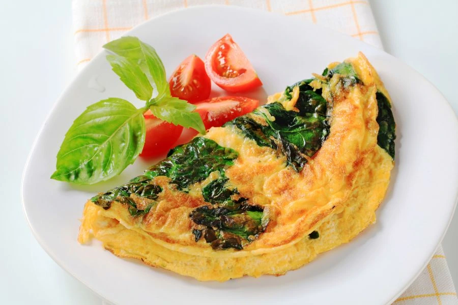 Omelete de Claras com Espinafre e Cogumelos servido em prato branco acompanhado de fatias de tomate fresco e folhas de manjericão, colocado sobre um pano branco com detalhes em xadrez. Opção de café da manhã saudável e nutritivo.
