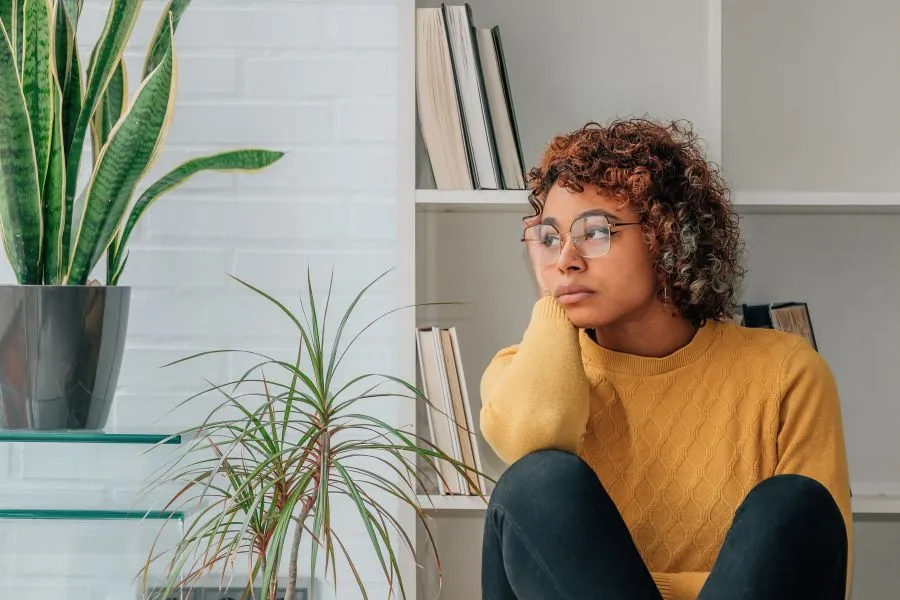 Mulher de óculos, com expressão pensativa, vestindo um suéter amarelo mostarda, sentada em um ambiente iluminado por luz natural. Ao fundo, uma estante branca com livros organizados, ao lado de plantas decorativas em vasos, incluindo um exemplar de espada-de-são-jorge.