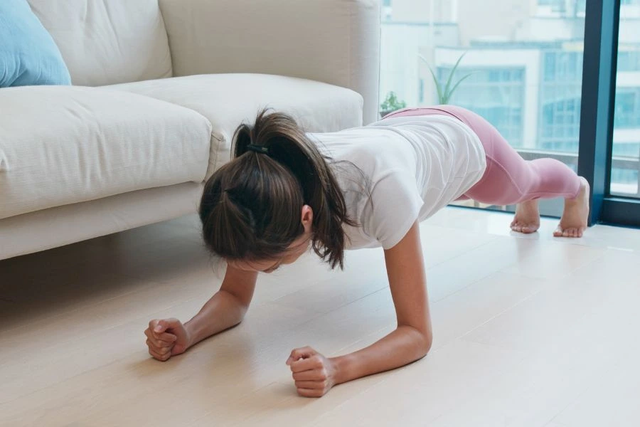 Mulher jovem realizando exercício de prancha no chão de madeira clara, vestindo camiseta branca e legging rosa. Ela mantém os antebraços apoiados no solo e a postura alinhada, demonstrando força e estabilidade. O ambiente caseiro, com sofá branco, janelas amplas e vista para edifícios, transmite conforto e dedicação à prática de exercícios físicos em casa.