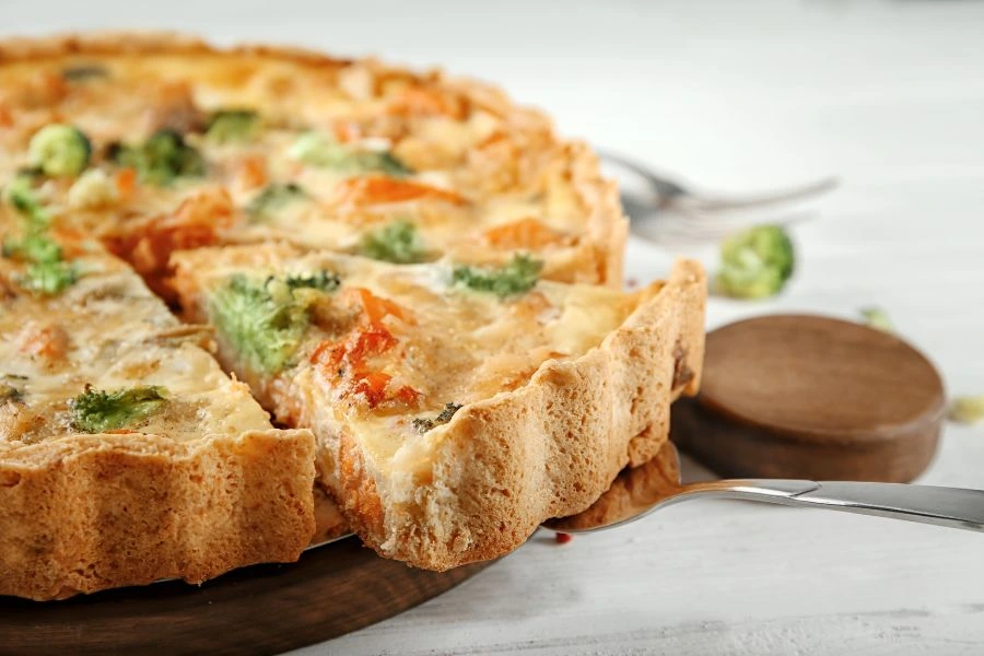 Quiche sem Massa de Brócolis e Bacon, servida sobre uma base de madeira. Uma fatia sendo retirada com uma espátula metálica, destacando a crocância da borda e a cremosidade do recheio. Prato ideal para um lanche ou refeição leve. Refeições Low Carb