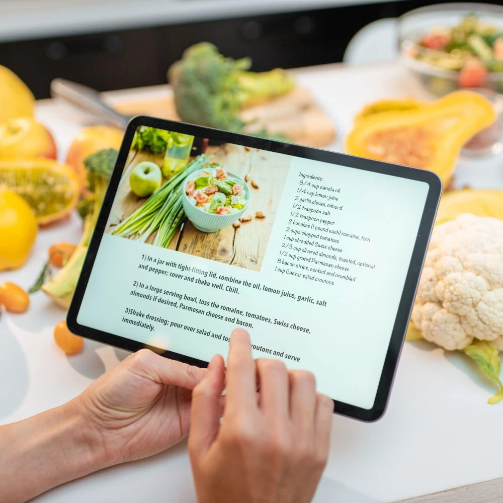 Pessoa segurando um tablet com uma receita de salada saudável exibida na tela. O tablet está rodeado por ingredientes frescos, como brócolis, abobrinha, pimentões, couve-flor e ervas verdes, dispostos sobre uma bancada branca. A composição destaca a preparação de refeições saudáveis em casa, com ênfase em alimentos naturais e estilo de vida equilibrado.