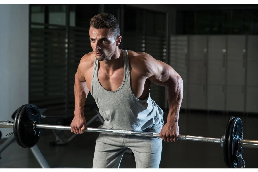 Homem jovem e fitness realizando um exercício de barra fixa com peso em uma academia bem equipada. Ele veste regata cinza e calça esportiva, exibindo músculos definidos enquanto mantém a postura inclinada durante o treino. Cena destaca intensidade, força muscular, e dedicação aos exercícios de musculação.