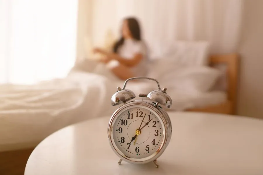 Despertador analógico em primeiro plano mostrando horário matinal, com pessoa desfocada ao fundo na cama, representando rotina de sono e importância de manter horários regulares para saúde menta