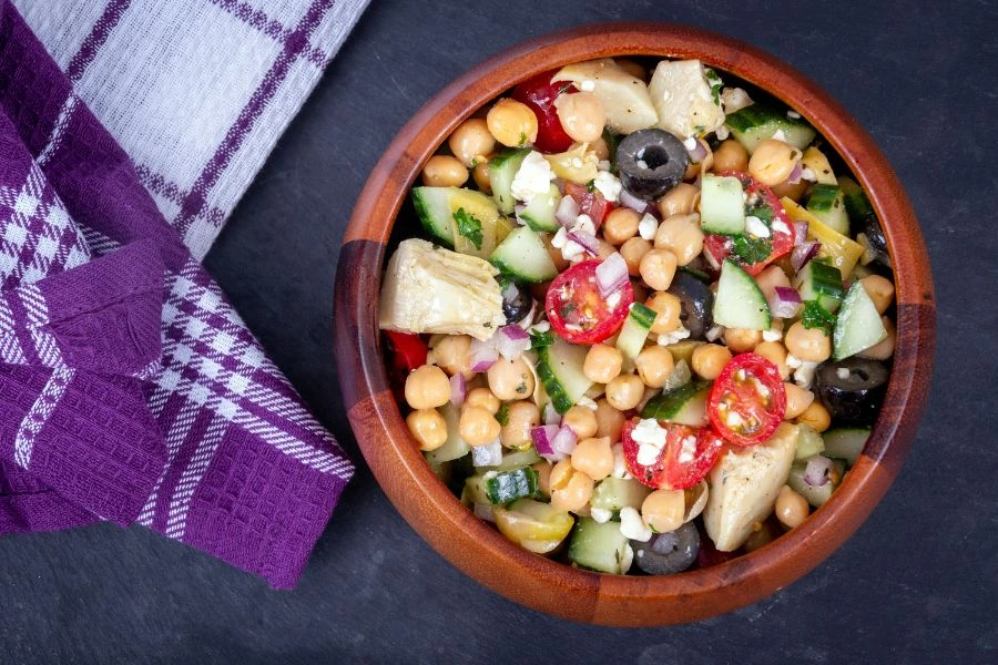 Salada mediterrânea de grão-de-bico em tigela de madeira, com tomate cereja, pepino, azeitonas pretas, cebola roxa, alcachofras e queijo feta, sobre fundo preto com pano roxo decorativo ao lado. Receita saudável e saborosa.