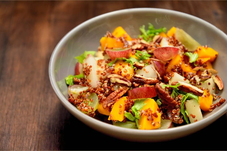 Tigela de Salada de Quinoa com Frutas e Nozes, servida em prato fundo cinza sobre uma mesa de madeira de tom escuro. Opção colorida, nutritiva e ideal para refeições leves.