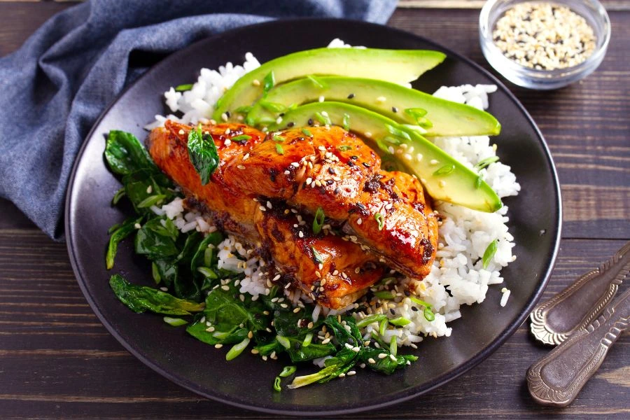 Prato de salmão grelhado com molho teriyaki, servido sobre uma cama de espinafre refogado e arroz branco, decorado com fatias frescas de abacate e sementes de gergelim. Refeição saudável e colorida apresentada em prato preto sobre uma mesa de madeira com guardanapo cinza ao lado.