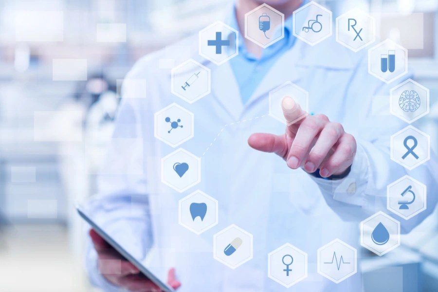 Médico vestindo jaleco branco interagindo com uma interface virtual de ícones relacionados à saúde, como coração, seringa, medicamentos e símbolo médico. Ele segura um tablet enquanto aponta para um dos ícones. O fundo desfocado mostra um ambiente clínico ou hospitalar, destacando tecnologia e inovação na área da saúde.