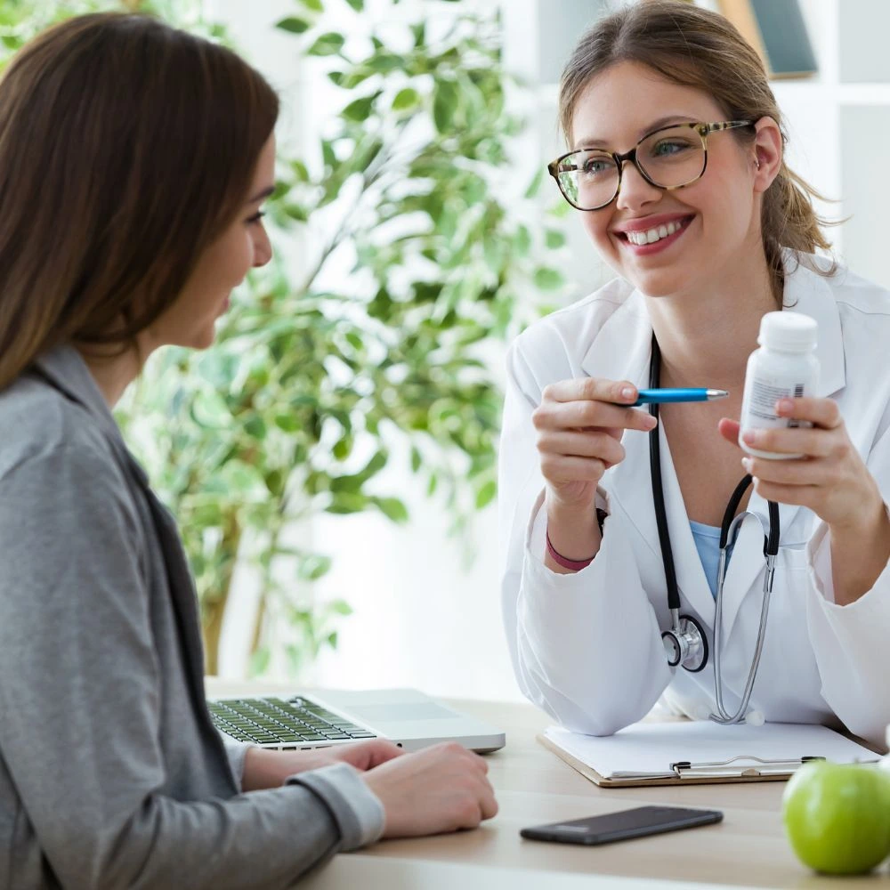 Profissional de saúde, vestindo jaleco branco e estetoscópio, sorrindo enquanto explica o uso de um frasco de medicamento para uma paciente em um consultório iluminado. A médica utiliza uma caneta para apontar o rótulo, enquanto a paciente ouve atentamente. Sobre a mesa estão um notebook, um smartphone e uma maçã verde, com uma planta ao fundo, criando um ambiente acolhedor e profissional, enfatizando cuidados com saúde e bem-estar.