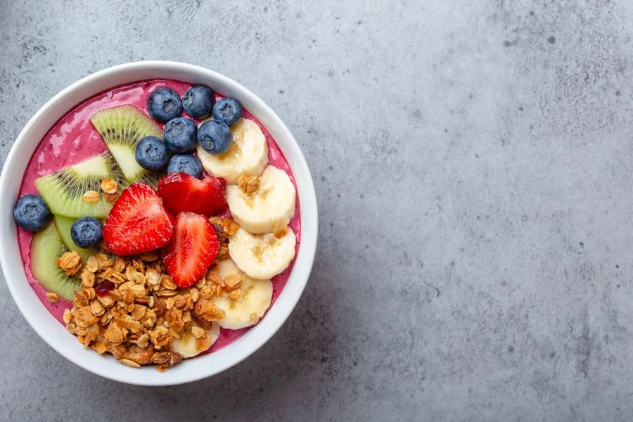 Tigela de smoothie de açaí com frutas vermelhas decorada com fatias de kiwi, morangos, bananas, mirtilos e granola crocante, sobre superfície de concreto cinza. Opção saudável para café da manhã ou lanche.