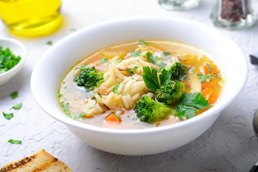 Sopa Rápida de Legumes com Macarrão Integral em tigela branca, contendo brócolis, cenoura, salsinha fresca e caldo quente. Prato servido em superfície clara com pão tostado ao lado. Opção reconfortante e saudável para dias frios.