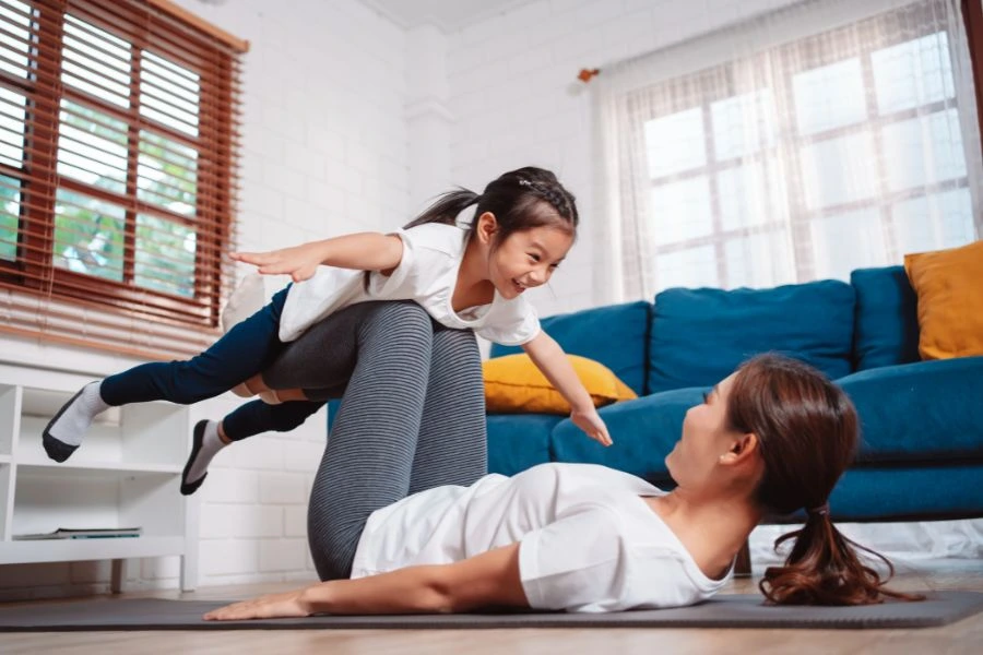 Mãe e filha brincando juntas em uma sala de estar iluminada e acolhedora. A mãe, deitada no chão, levanta a filha com as pernas enquanto a menina estende os braços simulando um avião, ambas sorrindo e aproveitando um momento alegre e afetuoso. Ao fundo, sofá azul com almofadas coloridas e janela trazendo luz natural, representando diversão em família e conexão emocional no ambiente doméstico.