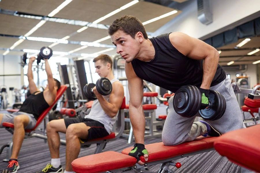 Homens em uma academia realizando exercícios de musculação com pesos. O primeiro homem em foco está inclinado sobre um banco, segurando um halter, enquanto outros dois, ao fundo, treinam em bancos ajustáveis. Cena representa treino de força, dedicação e estilo de vida saudável em um ambiente moderno de academia.