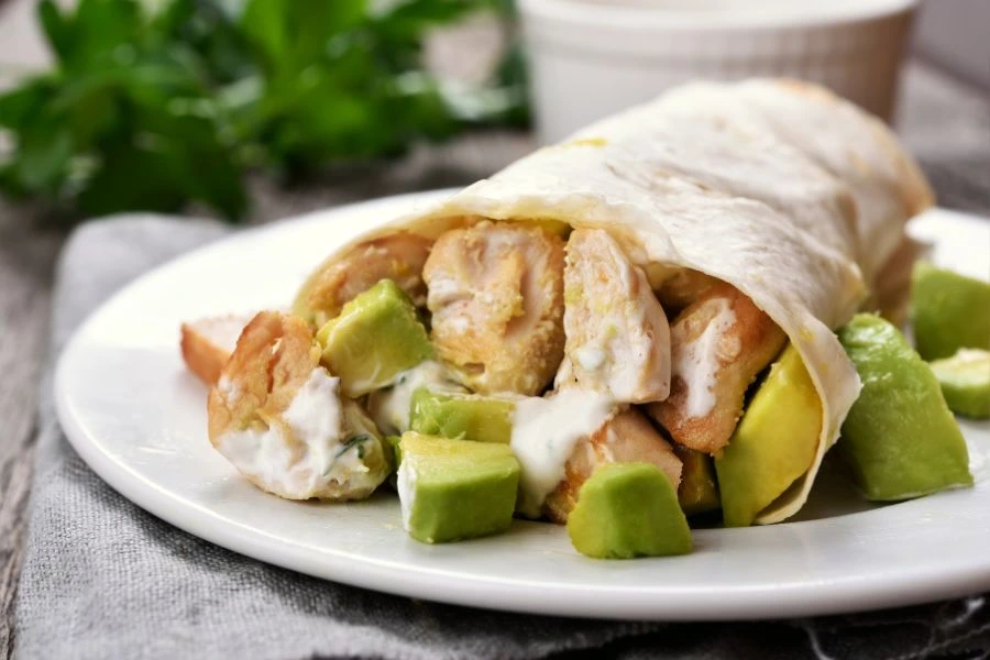 Wrap com pedaços de frango dourado, cubos de abacate fresco e molho cremoso, servido em um prato branco sobre uma mesa rústica com fundo desfocado de folhas verdes. Opção saborosa e saudável para refeições rápidas.
